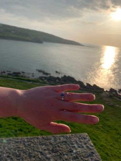 Platinum engagement ring with an oval sapphire and heirloom diamonds set either side with sunset in behind over Porthmeor beach